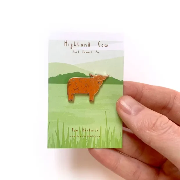 Highland Cow enamel pin badge, the cow is an orange brown colour and the pin is on a backing card of grass and hills.