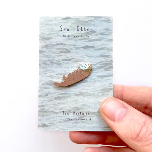Sea Otter enamel pin badge, the otter is brown and is on a decorative backing card which looks like water.